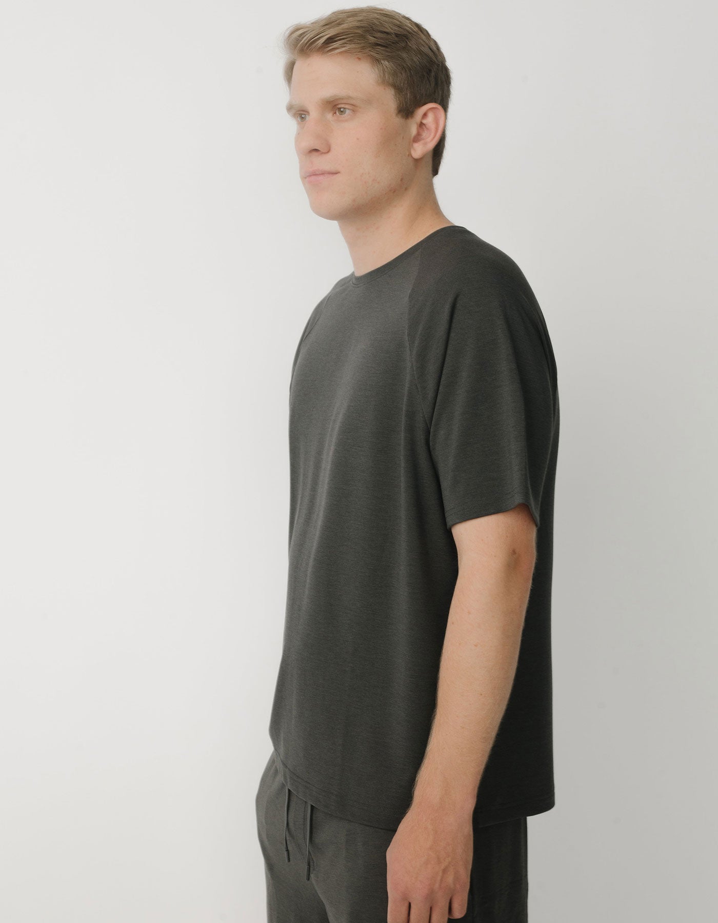 A person with short blonde hair stands in profile against a plain white background, wearing the Cozy Earth Men's Brushed Bamboo Raglan Tee - Last Chance in dark gray.