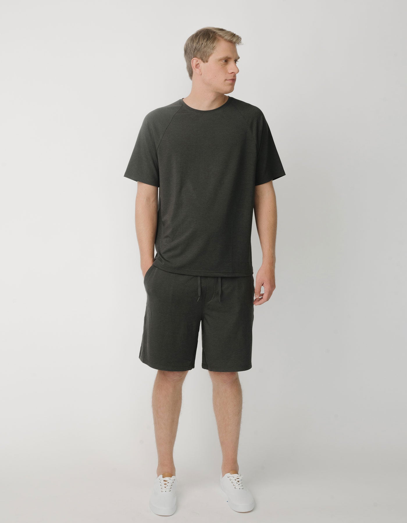 A man with light hair stands against a white background, wearing Cozy Earth’s Men's Brushed Bamboo Raglan Tee – Last Chance in dark gray with matching shorts and white sneakers. He is looking right, hands in his pockets.