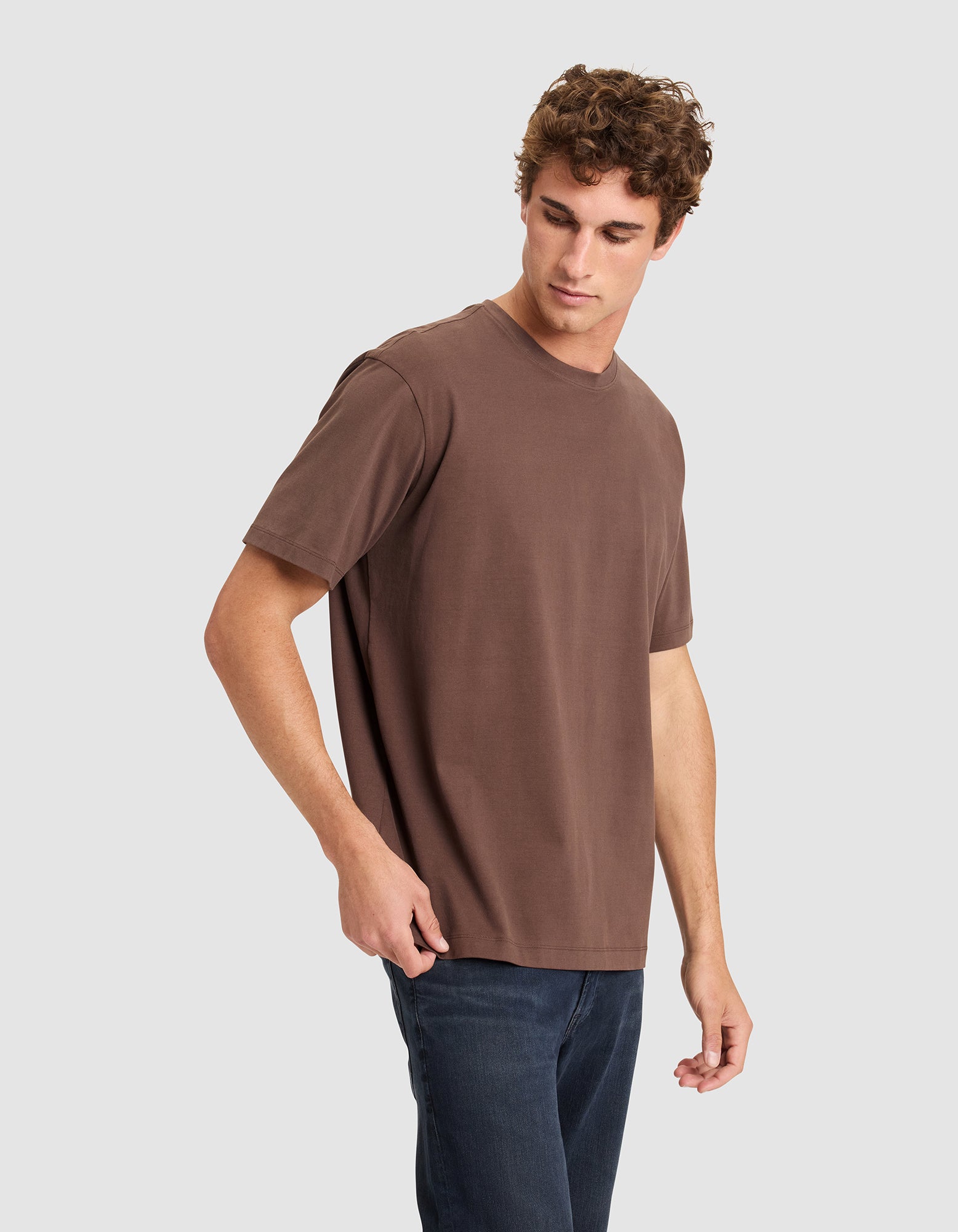 A young man with curly hair models the Cozy Earth Men's All Day Tee in brown and dark jeans, standing against a plain light gray background and looking down to his right.