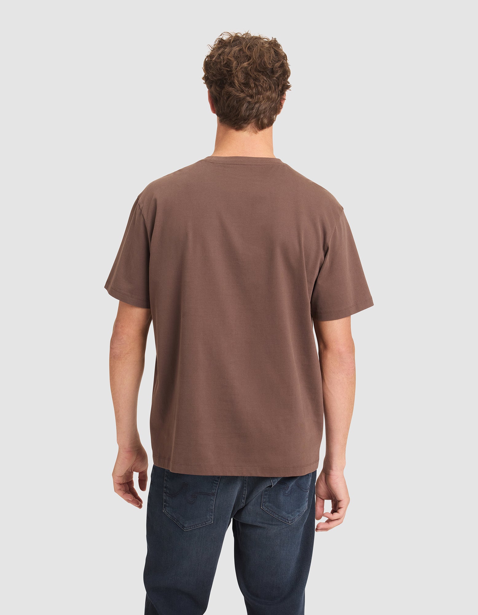 A man with short curly hair, seen from behind, wears a Cozy Earth Men's All Day Tee in brown and dark blue jeans against a plain light background. 