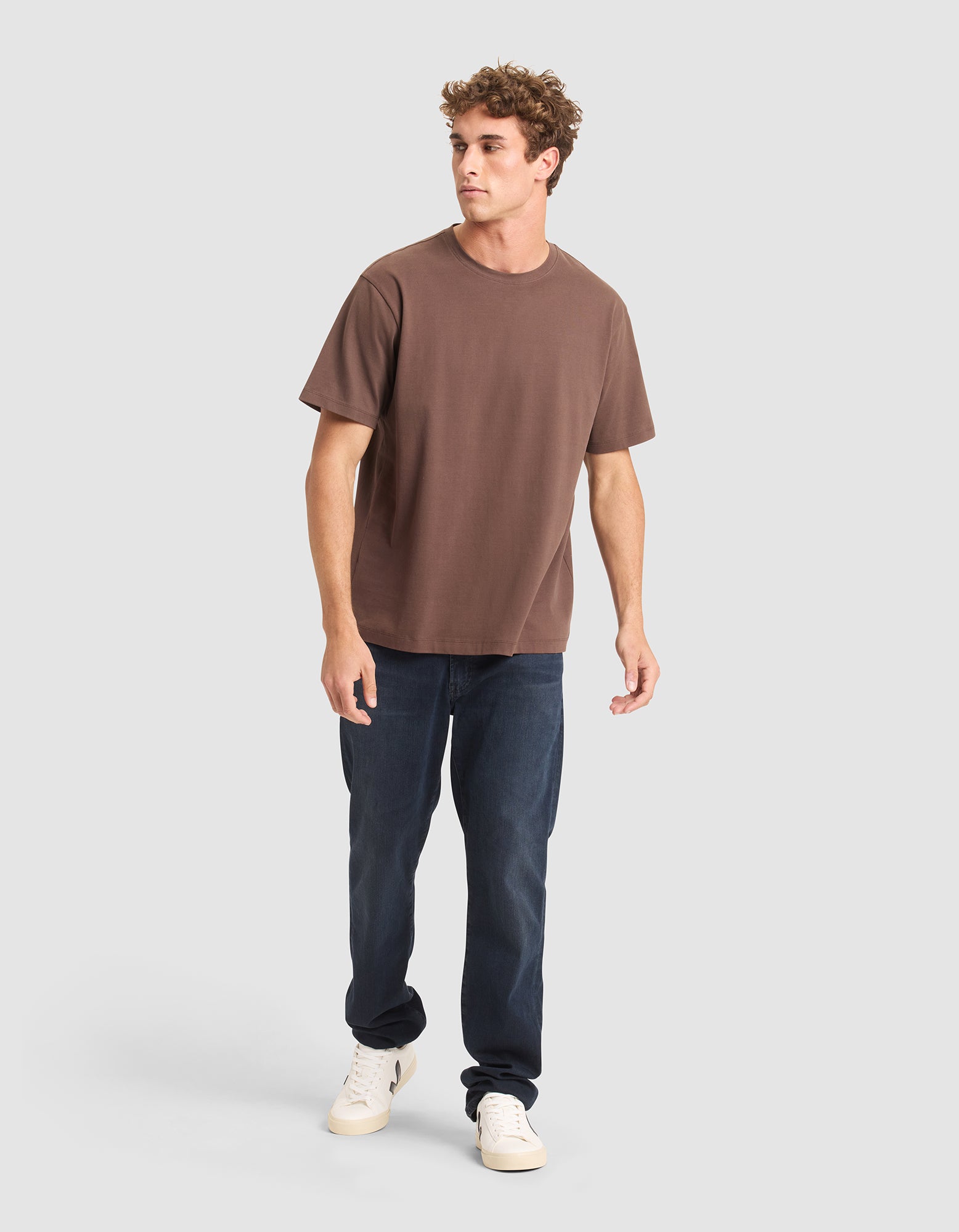 A young man stands on a light gray background wearing the Cozy Earth Men's All Day Tee in brown, paired with dark blue jeans and white sneakers. He looks slightly to the side, one leg forward as if walking. 