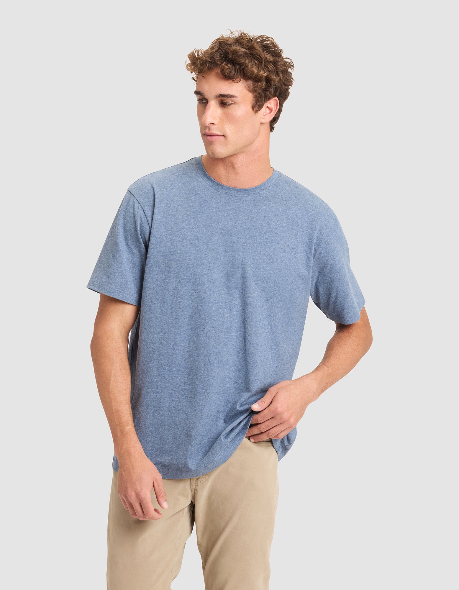 A young man with curly brown hair models the Cozy Earth Men's All Day Tee in light blue, paired with beige pants. He stands against a plain light gray background, gazing left and holding the hem of his shirt. 