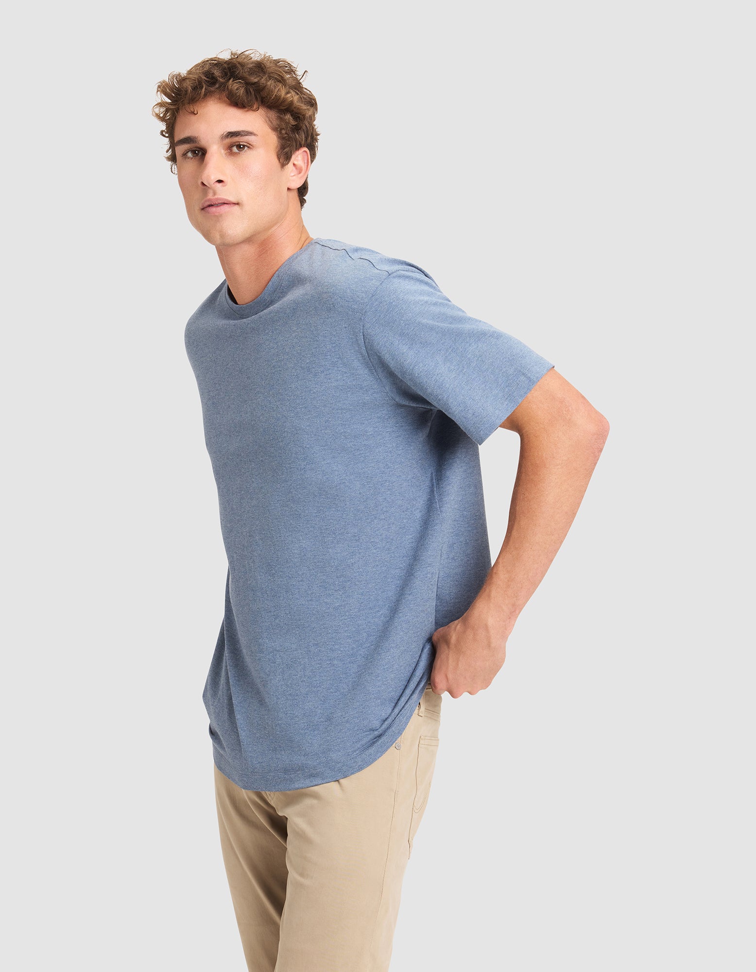 A young man with curly hair models the Cozy Earth Men's All Day Tee in light blue and beige pants, standing sideways with one hand in his pocket against a plain light gray background. 