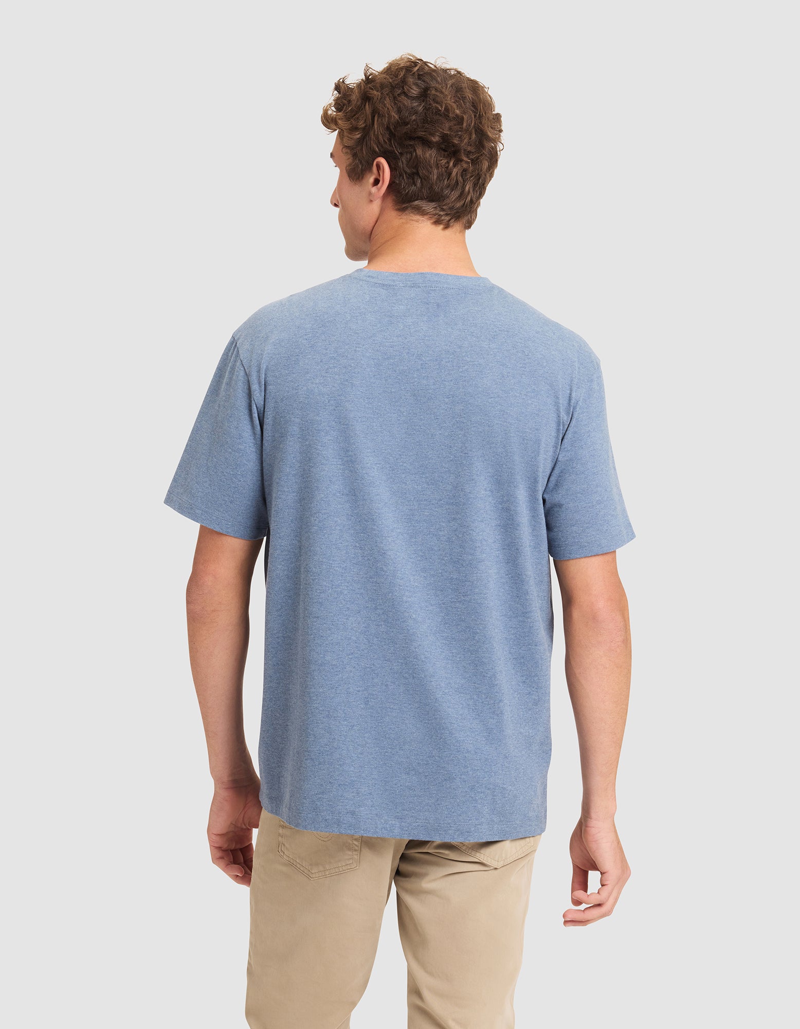 A man with short curly hair is standing and facing away, wearing the Cozy Earth Men's All Day Tee in blue and beige pants against a plain light background. 