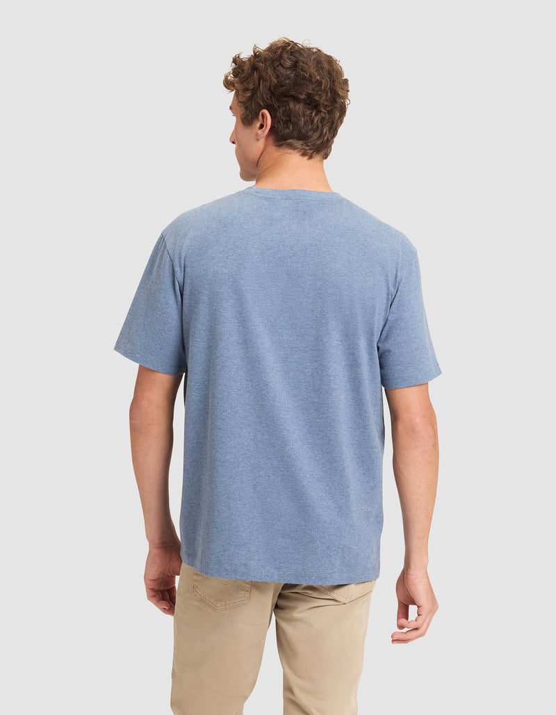 A man with short curly hair is standing and facing away, wearing the Cozy Earth Men's All Day Tee in blue and beige pants against a plain light background.