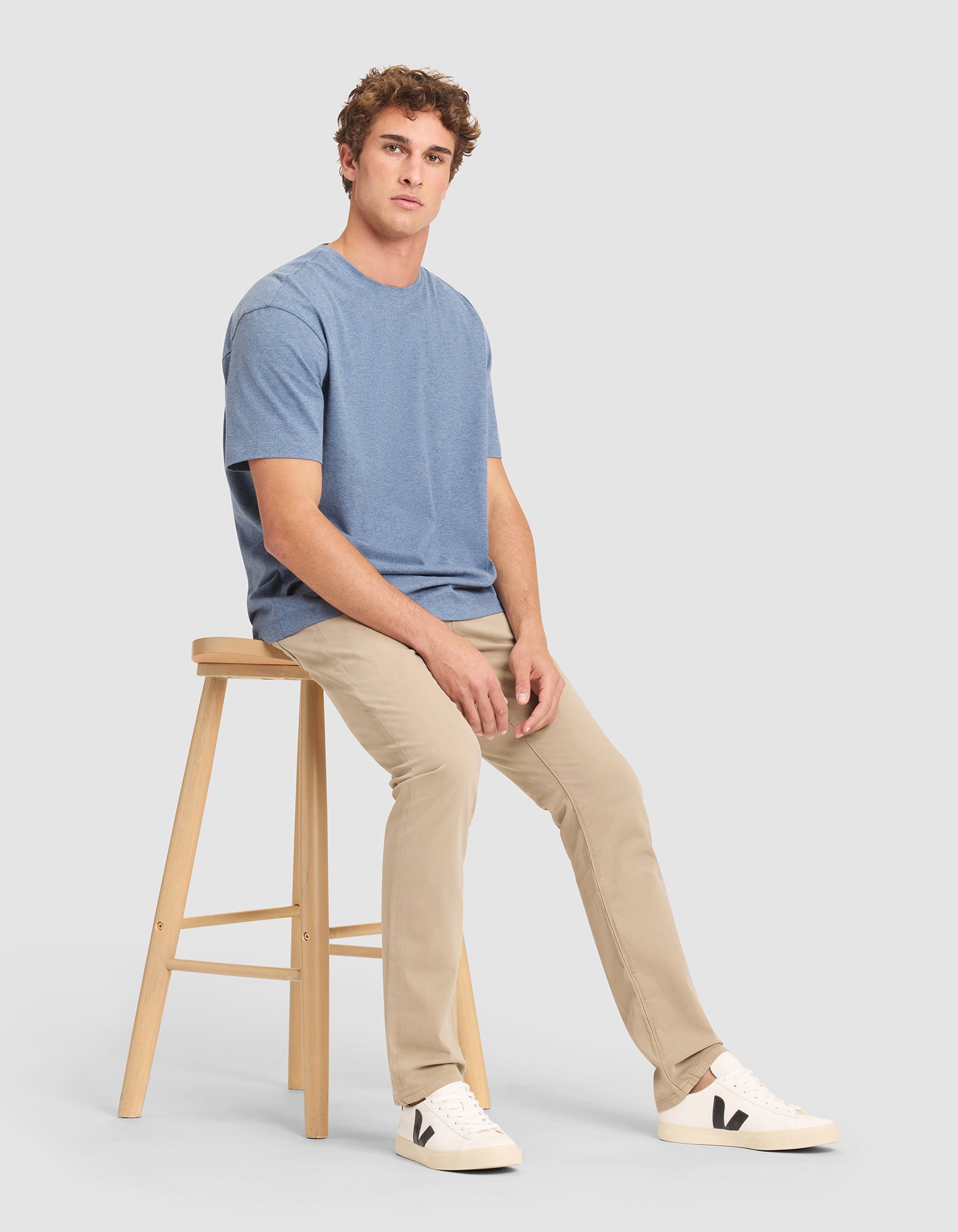 A man in the Cozy Earth Men's All Day Tee sits on a wooden stool against a plain light gray background. 
