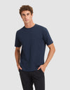 A young man with curly brown hair stands with his hands in his pockets against a light gray background, wearing the Cozy Earth Men's All Day Tee in navy blue and black pants. |Color:Eclipse