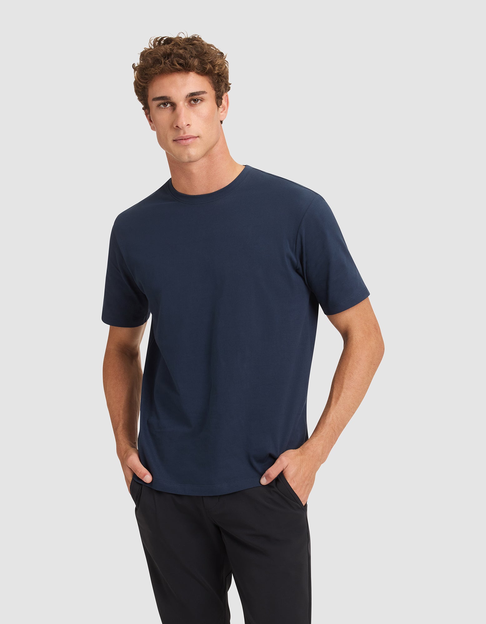 A young man with curly brown hair stands with his hands in his pockets against a light gray background, wearing the Cozy Earth Men's All Day Tee in navy blue and black pants. 