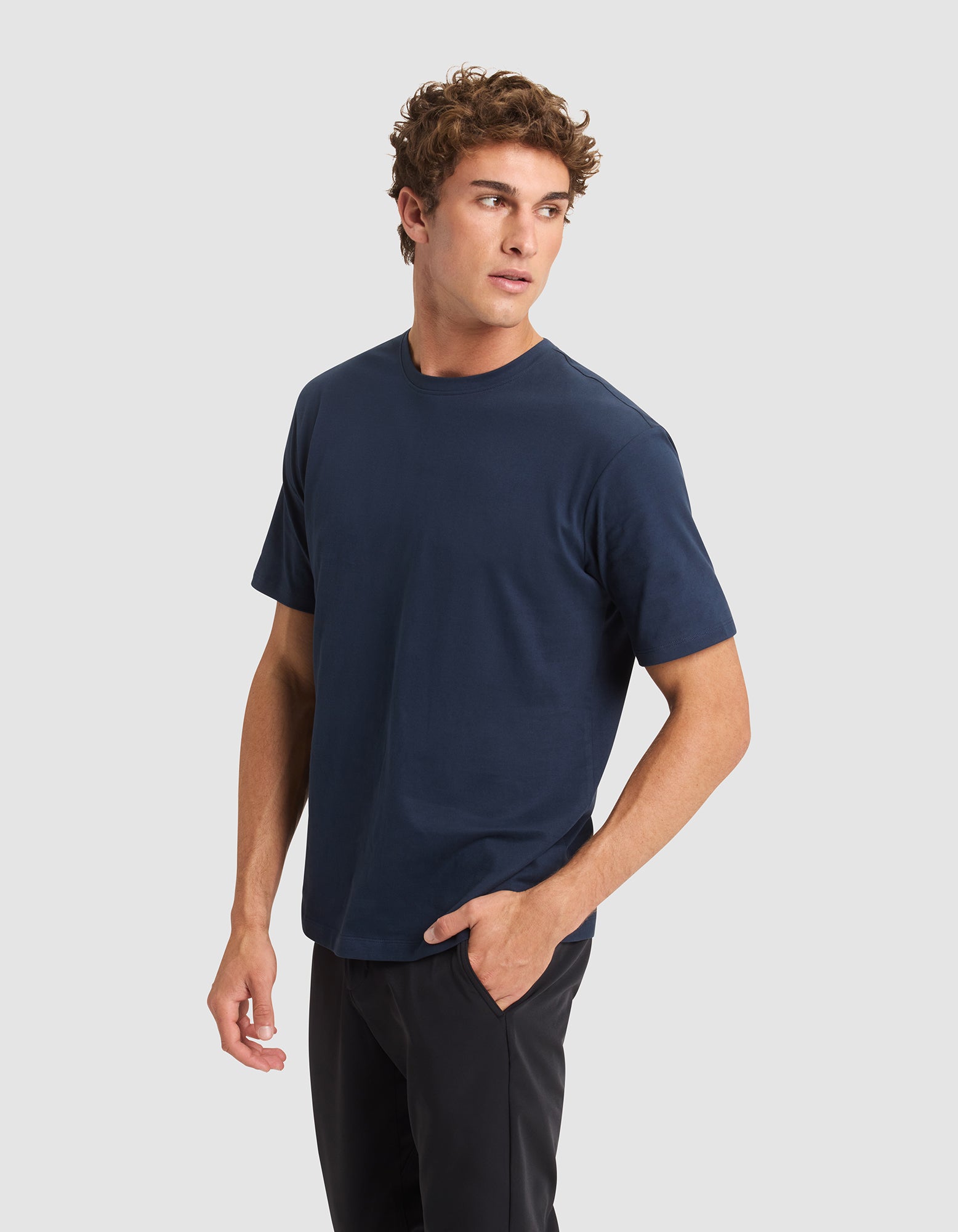A man with curly brown hair wears Cozy Earth's Men's All Day Tee in navy blue and black pants, standing against a light gray background and looking left with one hand in his pocket. 