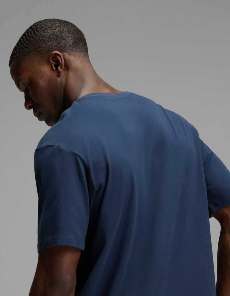A man with short hair is shown from the back and side, looking downward against a plain gray background while wearing the Cozy Earth Men’s All Day Tee in navy blue.