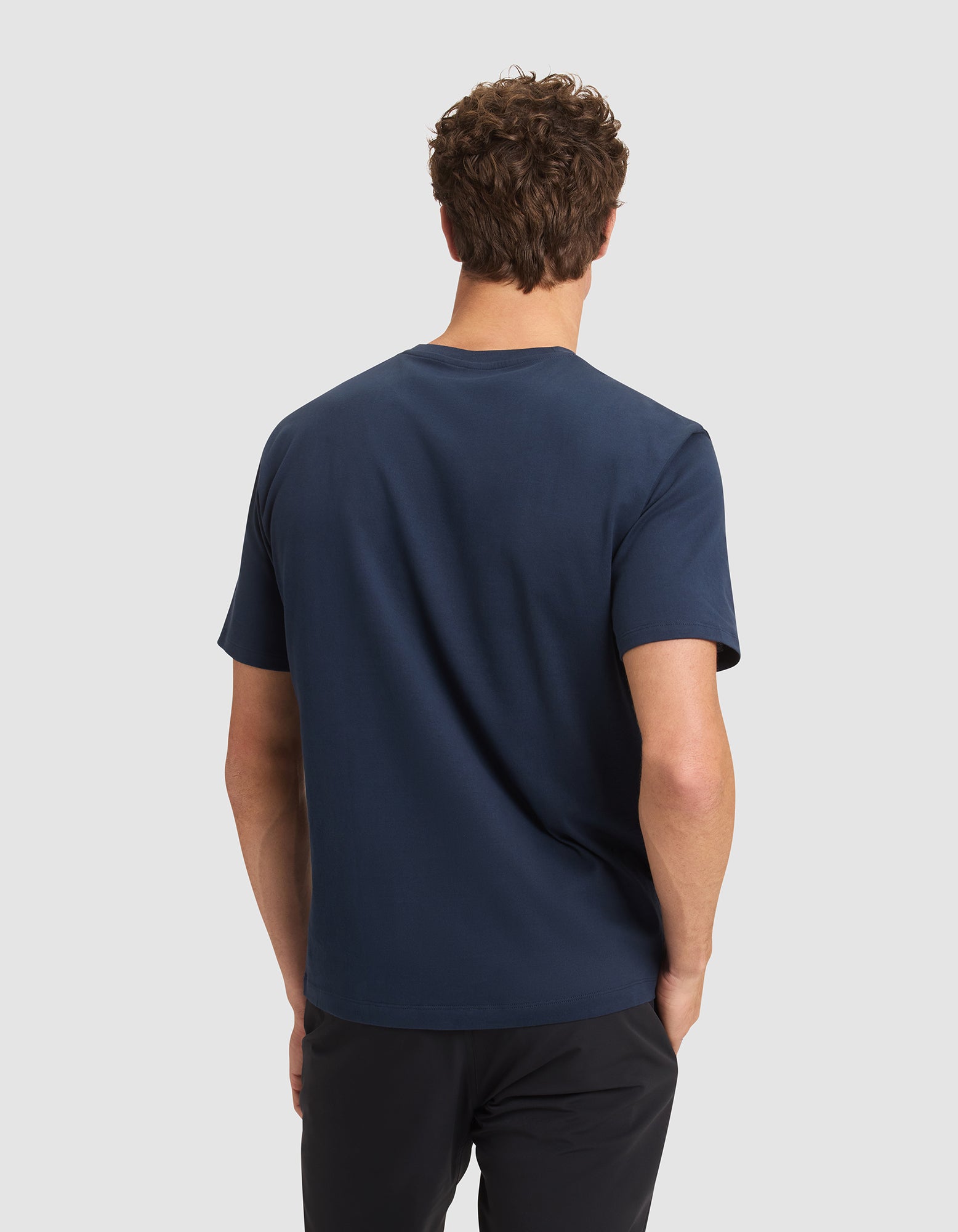 A man with short curly hair stands with his back to the camera, wearing the Cozy Earth Men's All Day Tee in navy blue and black pants against a light gray background. 