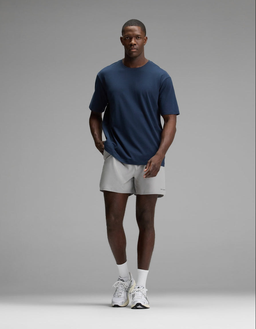A man stands against a plain gray background wearing the Cozy Earth Men’s All Day Tee in navy blue, paired with light gray shorts, white socks, and white sneakers, with his hands in his pockets.