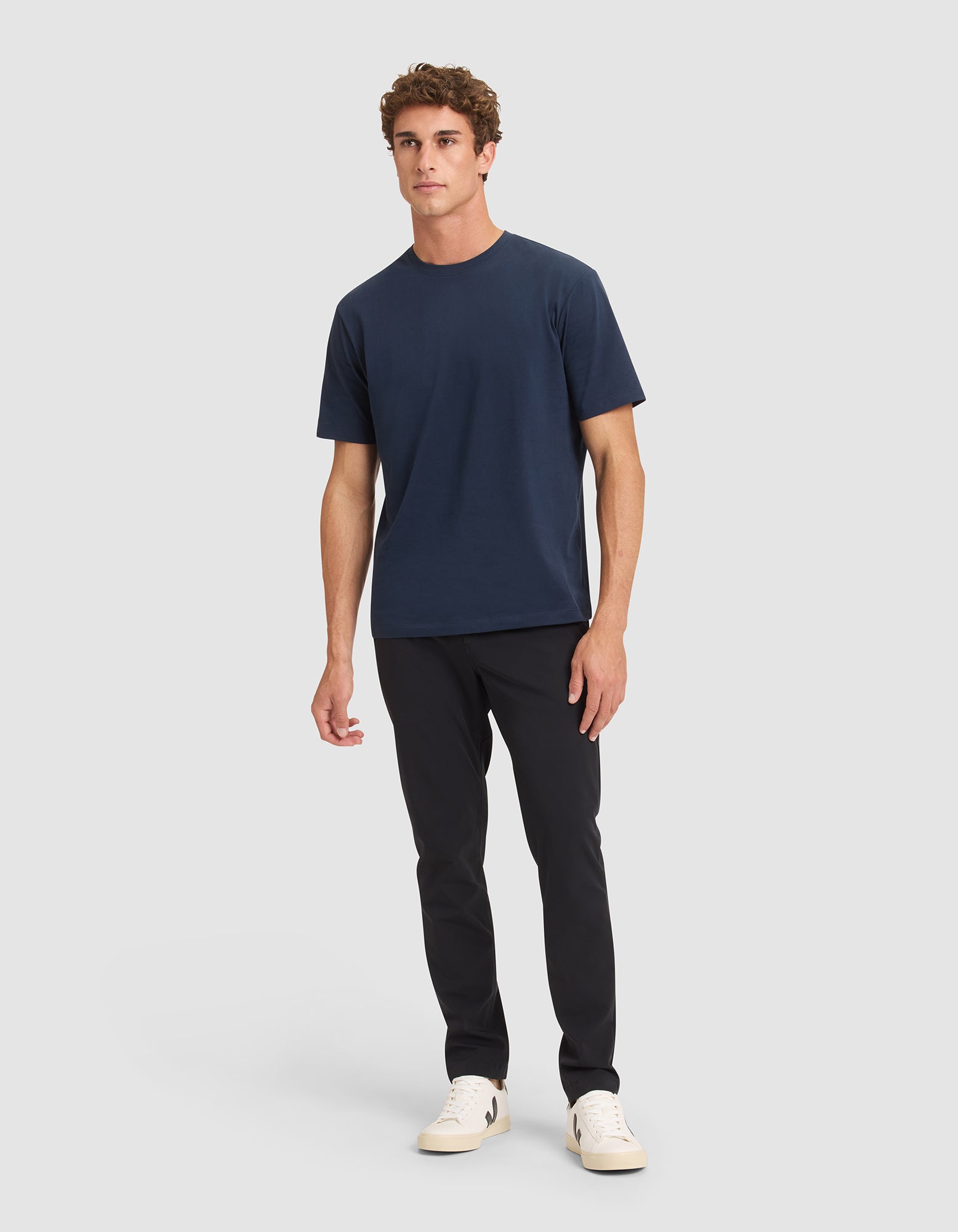 A young man stands against a plain light background wearing the Cozy Earth Men's All Day Tee in navy blue, paired with black pants and white sneakers. He faces forward with a relaxed posture and neutral expression. 