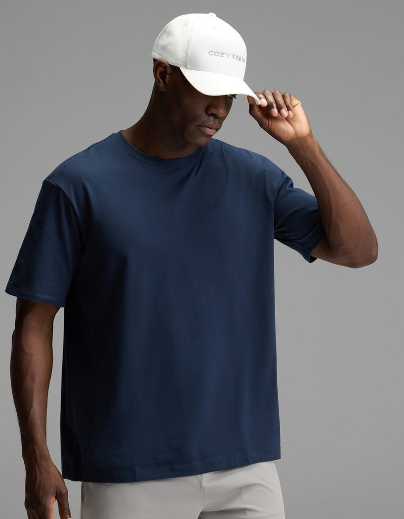 A man wearing the Cozy Earth Men’s All Day Tee in navy blue, paired with light gray pants and a white “COZY FARM” baseball cap, looks down and adjusts his cap against a plain gray background.