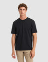 A young man with curly brown hair models the Cozy Earth Men's All Day Tee in black with beige pants, standing against a light gray background and looking straight at the camera with a neutral expression. |Color:Jet Black