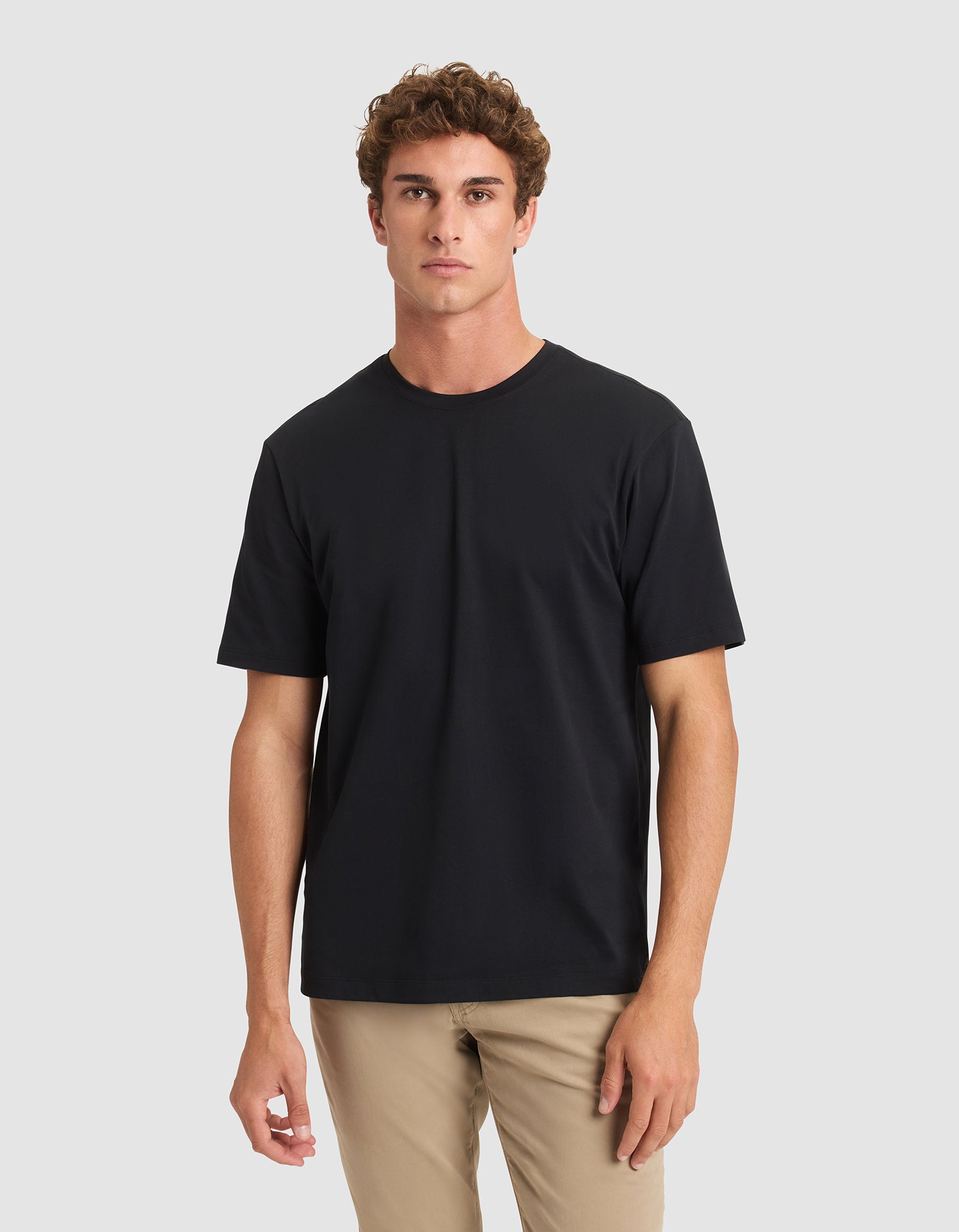 A young man with curly brown hair models the Cozy Earth Men's All Day Tee in black with beige pants, standing against a light gray background and looking straight at the camera with a neutral expression. 