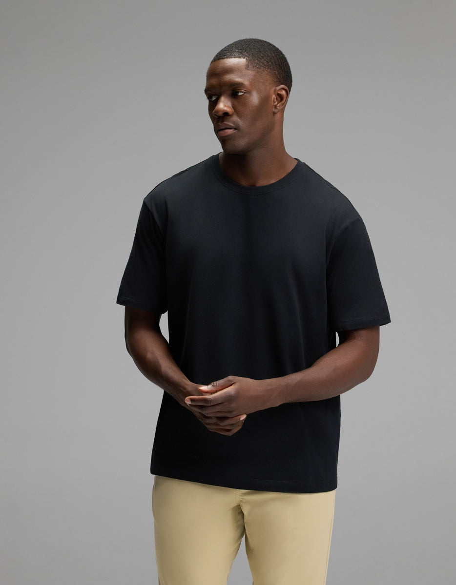 A man wearing the Cozy Earth Men’s All Day Tee in Jet Black and beige pants stands against a gray background, looking to his left with his hands loosely clasped.|Color:Jet Black