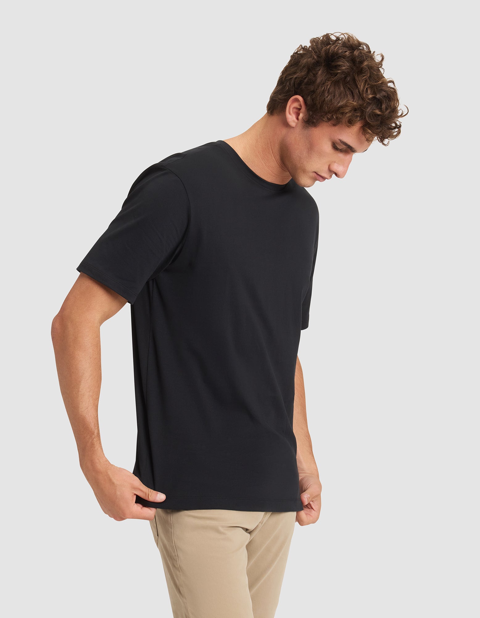 A young man with curly brown hair wears the Cozy Earth Men's All Day Tee in plain black and beige pants, standing against a light gray background and looking downward with his hands at his sides. 