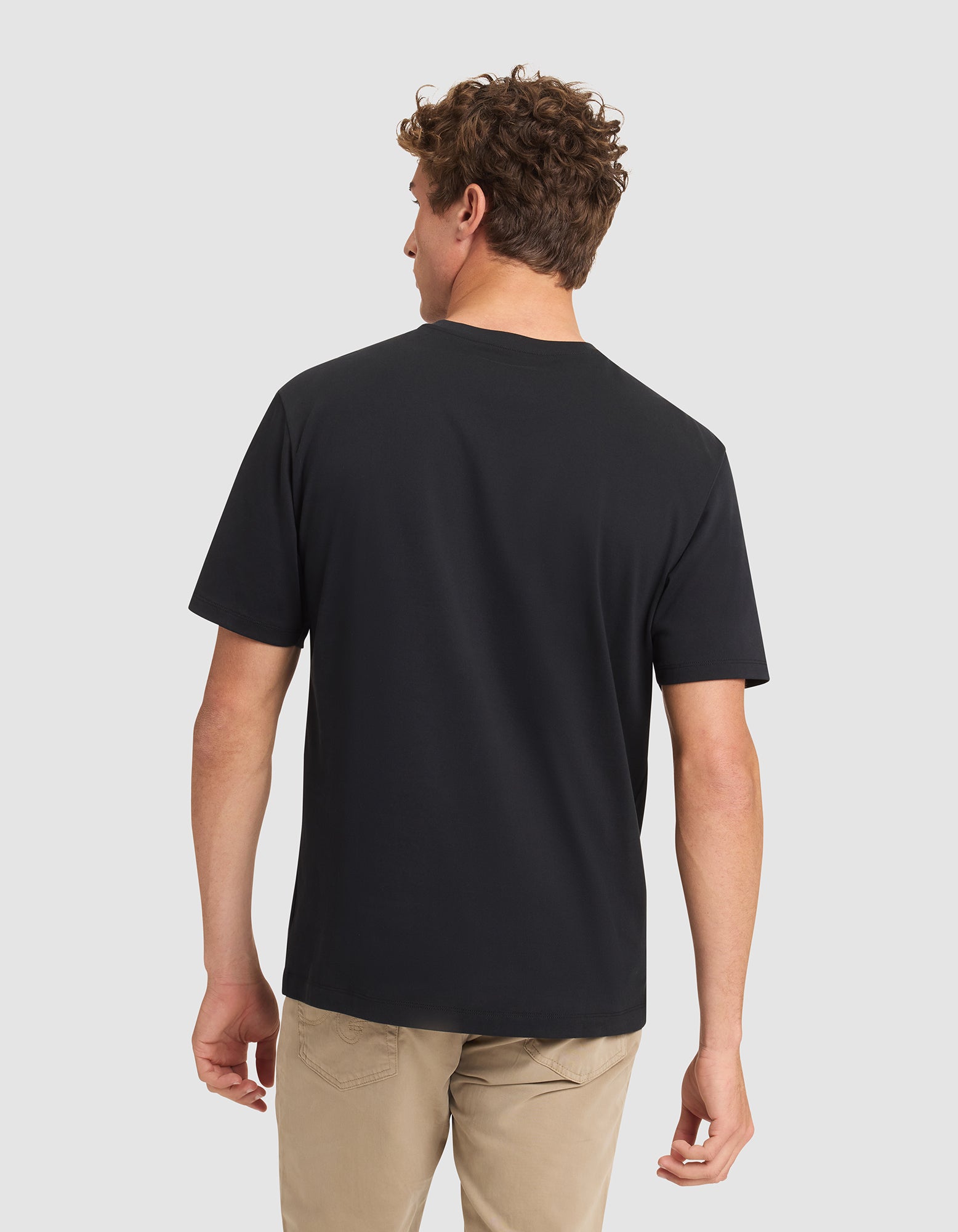 A man with short curly hair stands with his back to the camera, wearing a Cozy Earth Men's All Day Tee in black and beige pants against a light gray background. 