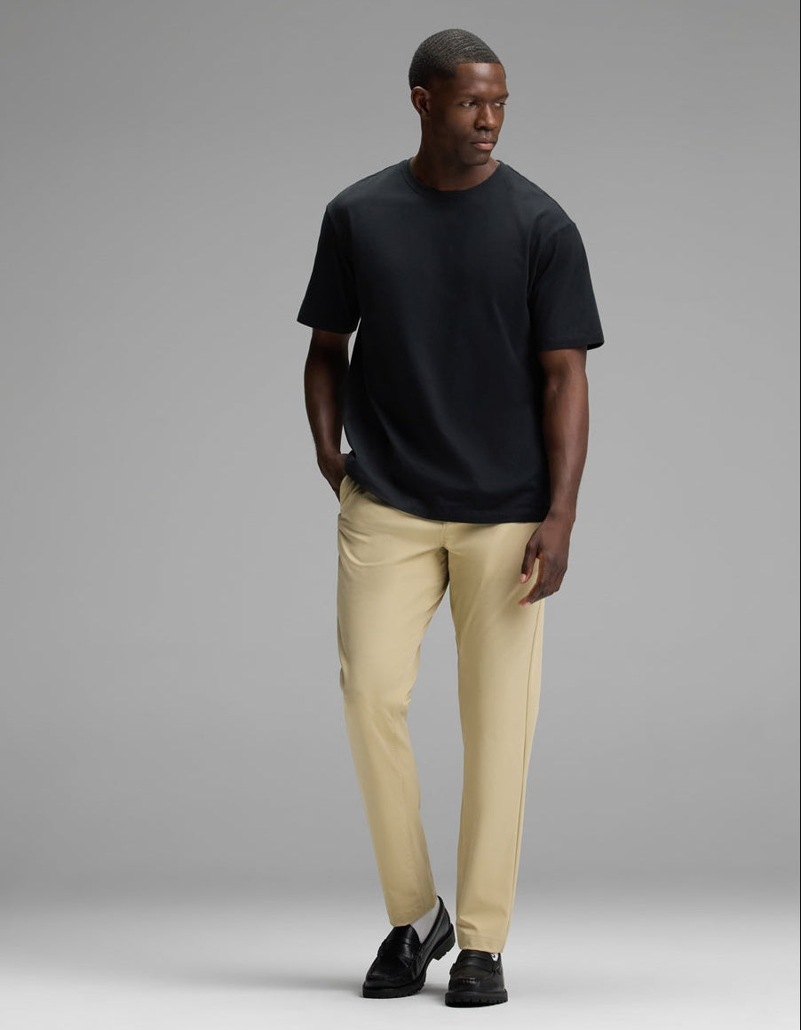 A man stands against a gray background, wearing Cozy Earth’s Men’s All Day Tee in black, paired with beige pants and black shoes. He has one hand in his pocket and is looking to his left.