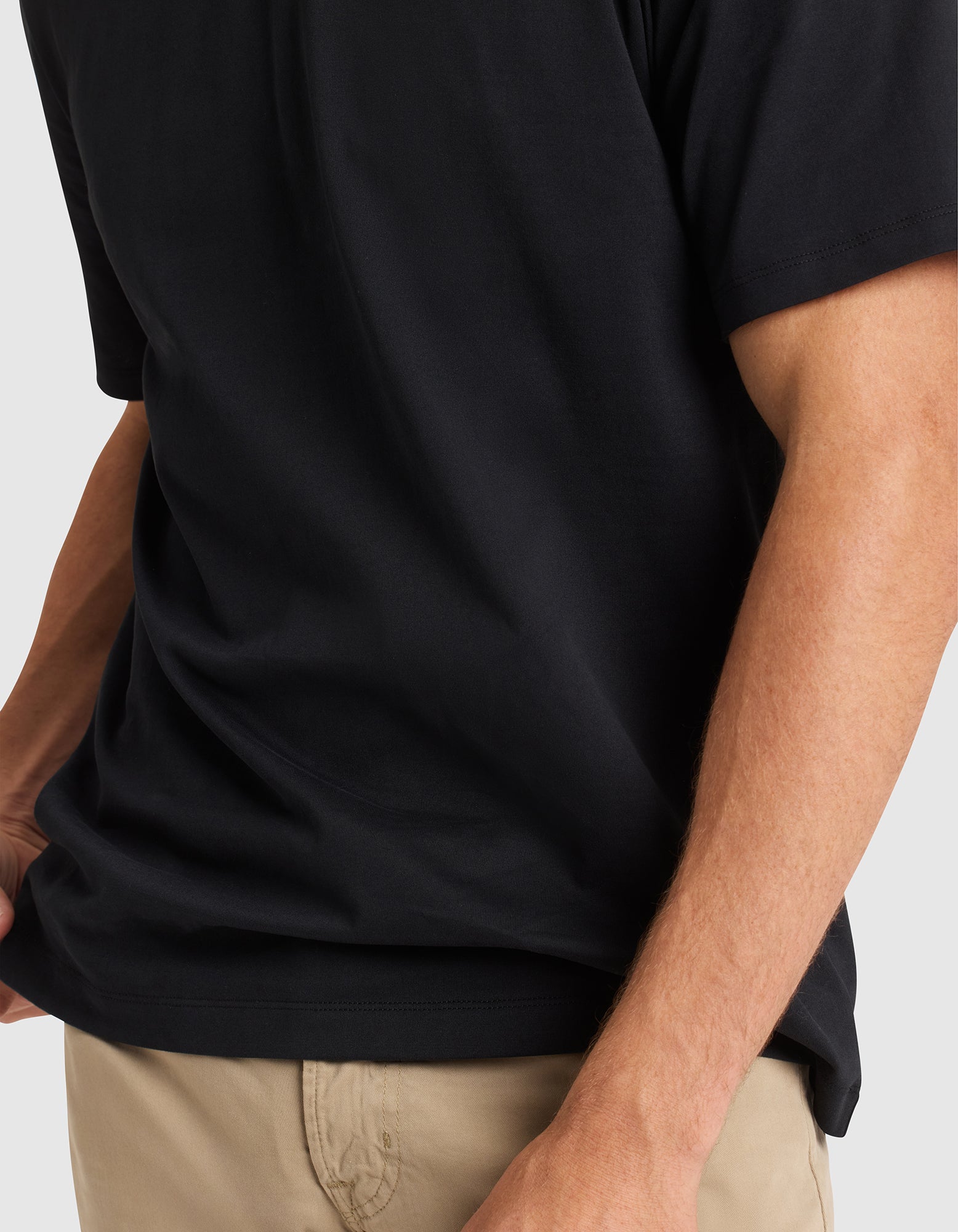 A man wears the Cozy Earth Men's All Day Tee in black and beige pants, shown waist to mid-arm with his left hand in his pocket. The background is plain and light-colored. 