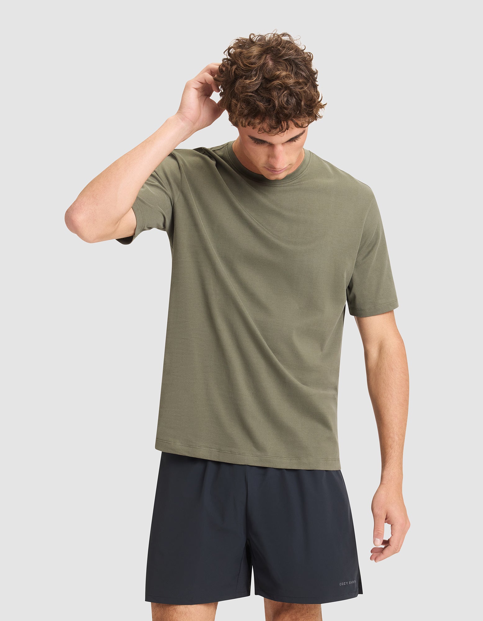 A man with curly brown hair wears the Cozy Earth Men's All Day Tee in olive green and black shorts, standing against a plain light gray background, looking down and touching the back of his head. |Color:Olive