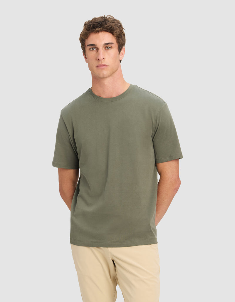 A young man with short curly brown hair models the Cozy Earth Men's All Day Tee in olive green, paired with beige pants, standing against a plain light gray background with his hands behind his back.