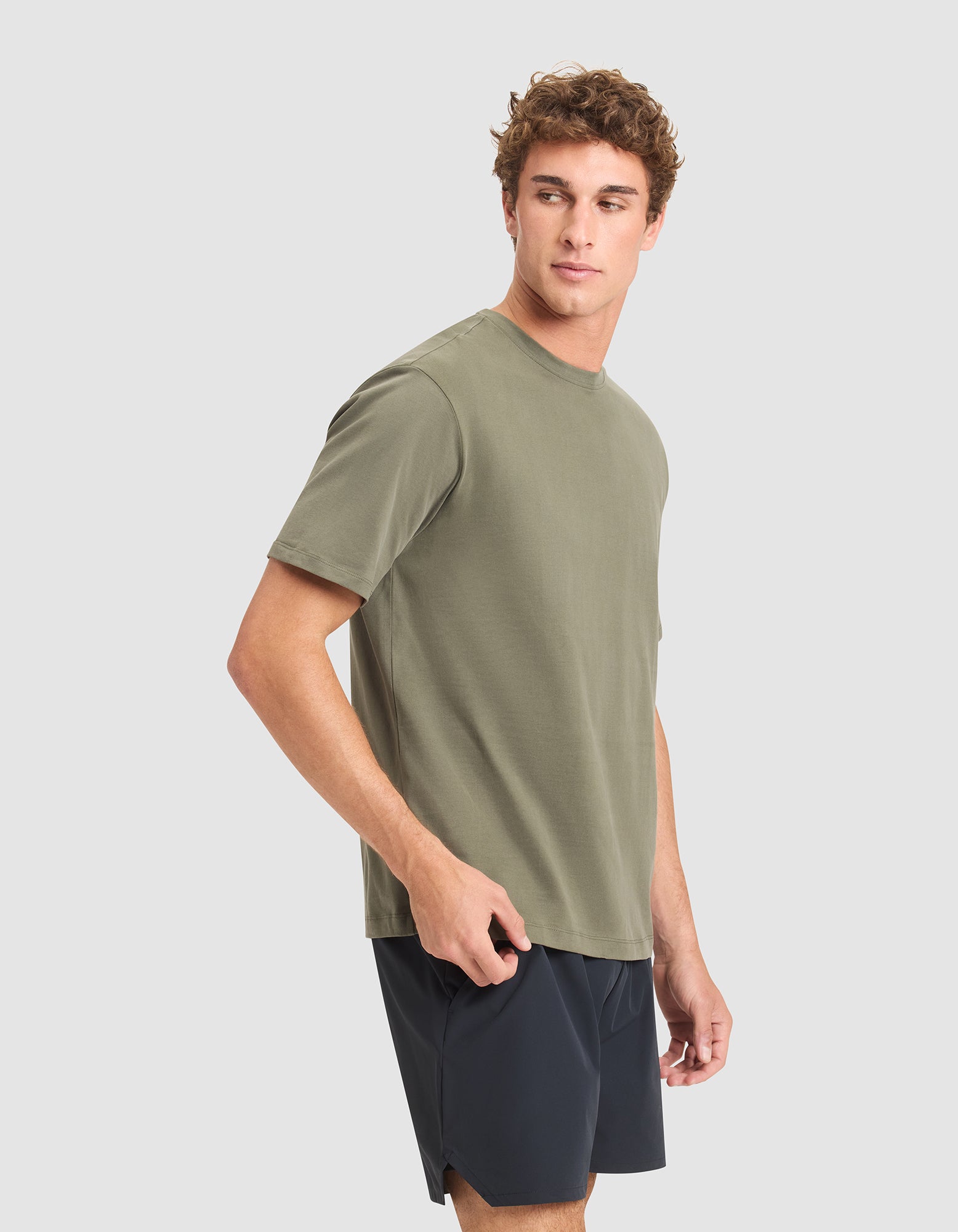 A young man with curly hair models the Cozy Earth Men's All Day Tee in olive green paired with black shorts, standing relaxed against a light gray background. 