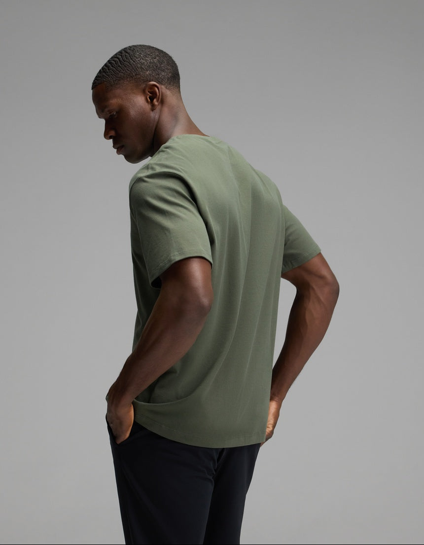 A man is seen from behind, wearing the Cozy Earth Men’s All Day Tee in olive green paired with black pants, standing against a plain gray background and looking slightly downward.|Color:Olive