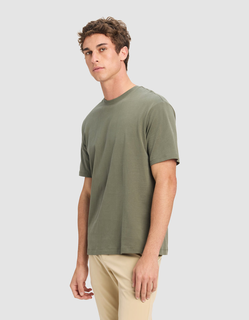 A man with short brown hair wears the Cozy Earth Men's All Day Tee in olive green and beige pants, standing against a plain light gray background and looking slightly to the side.