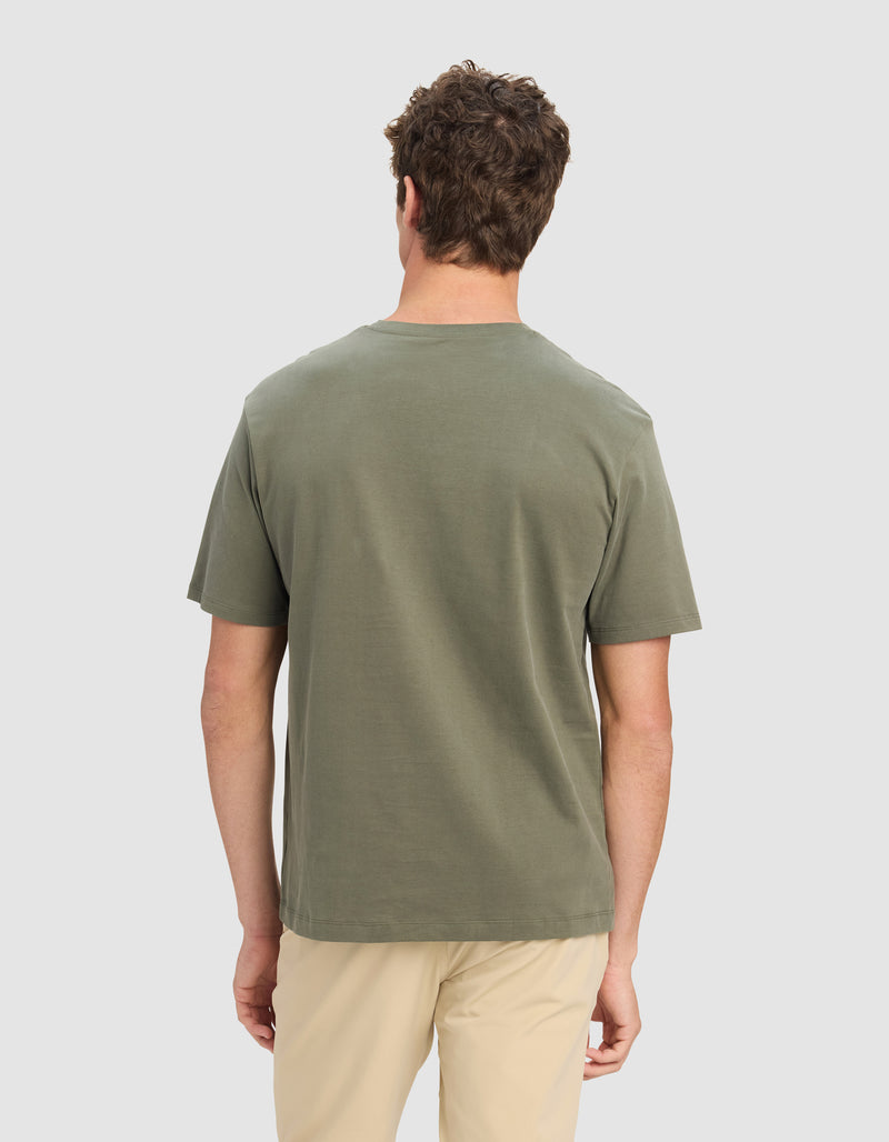 A man with short curly hair stands with his back to the camera, wearing the Cozy Earth Men's All Day Tee in olive green and light beige pants, set against a plain white background.