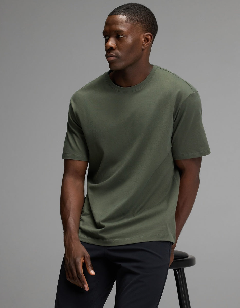 A man wearing the Cozy Earth Men’s All Day Tee in olive and black pants sits on a black stool against a plain gray background, looking to his left with a neutral expression.|Color:Olive