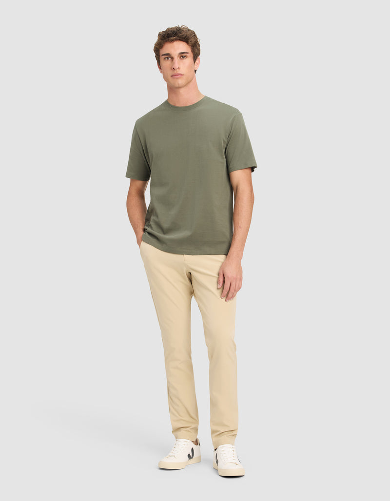 A young man with short brown hair stands against a plain background, hands in his pockets, wearing the Cozy Earth Men's All Day Tee in olive green, paired with beige pants and white sneakers.