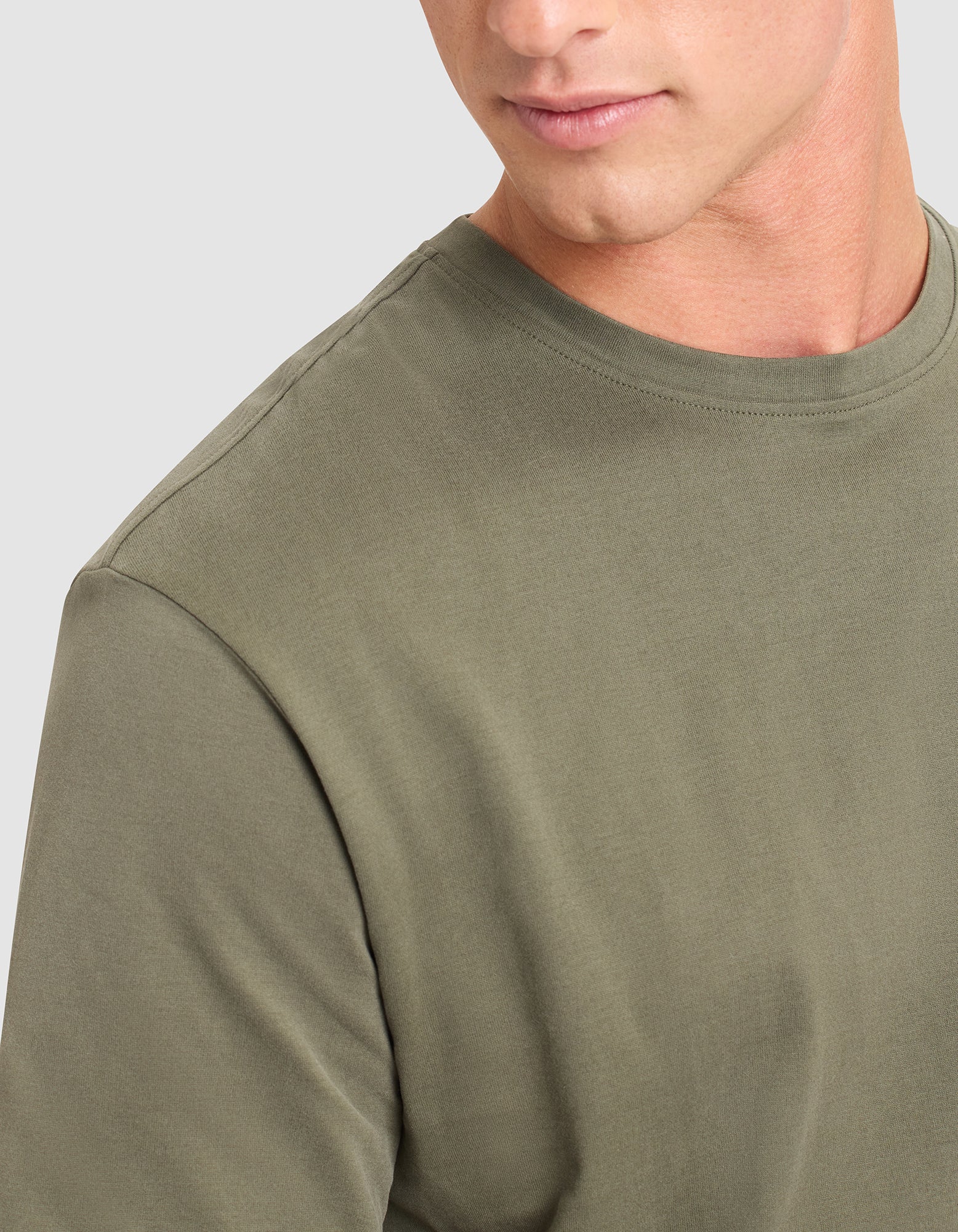 A young man is shown from the shoulders up, looking slightly downward and to his left, wearing the Cozy Earth Men's All Day Tee in plain olive green against a light gray background. 
