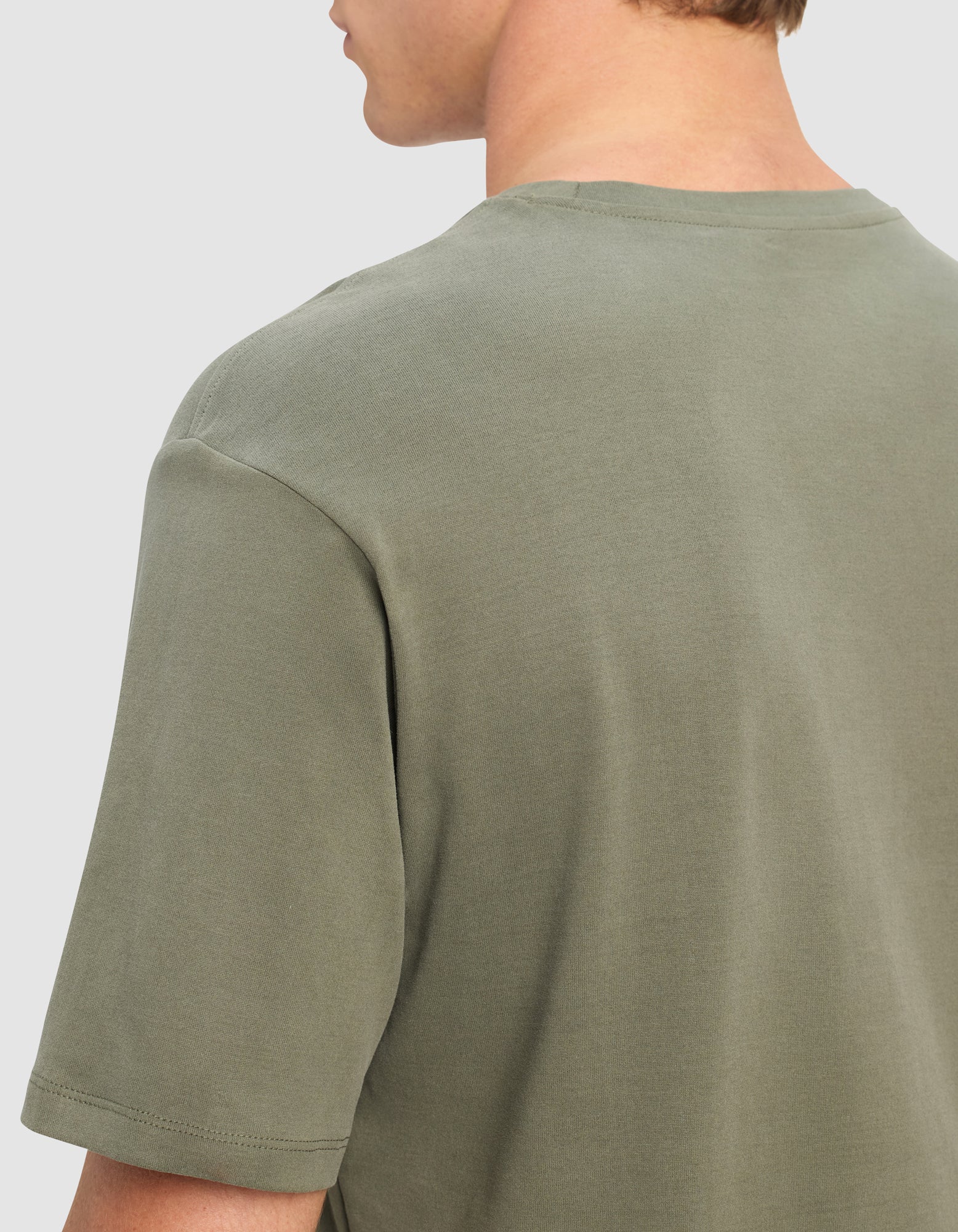 A close-up of a person wearing the Cozy Earth Men's All Day Tee in sage green, shown from the back and slightly to the side. The short-sleeve shirt is plain with a classic crew neckline, and the person's face is not visible.