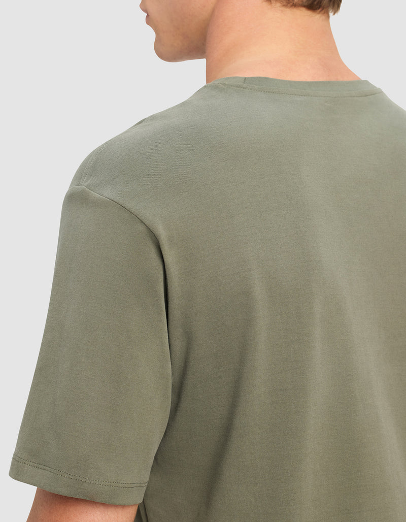 A close-up of a person wearing the Cozy Earth Men's All Day Tee in sage green, shown from the back and slightly to the side. The short-sleeve shirt is plain with a classic crew neckline, and the person's face is not visible.