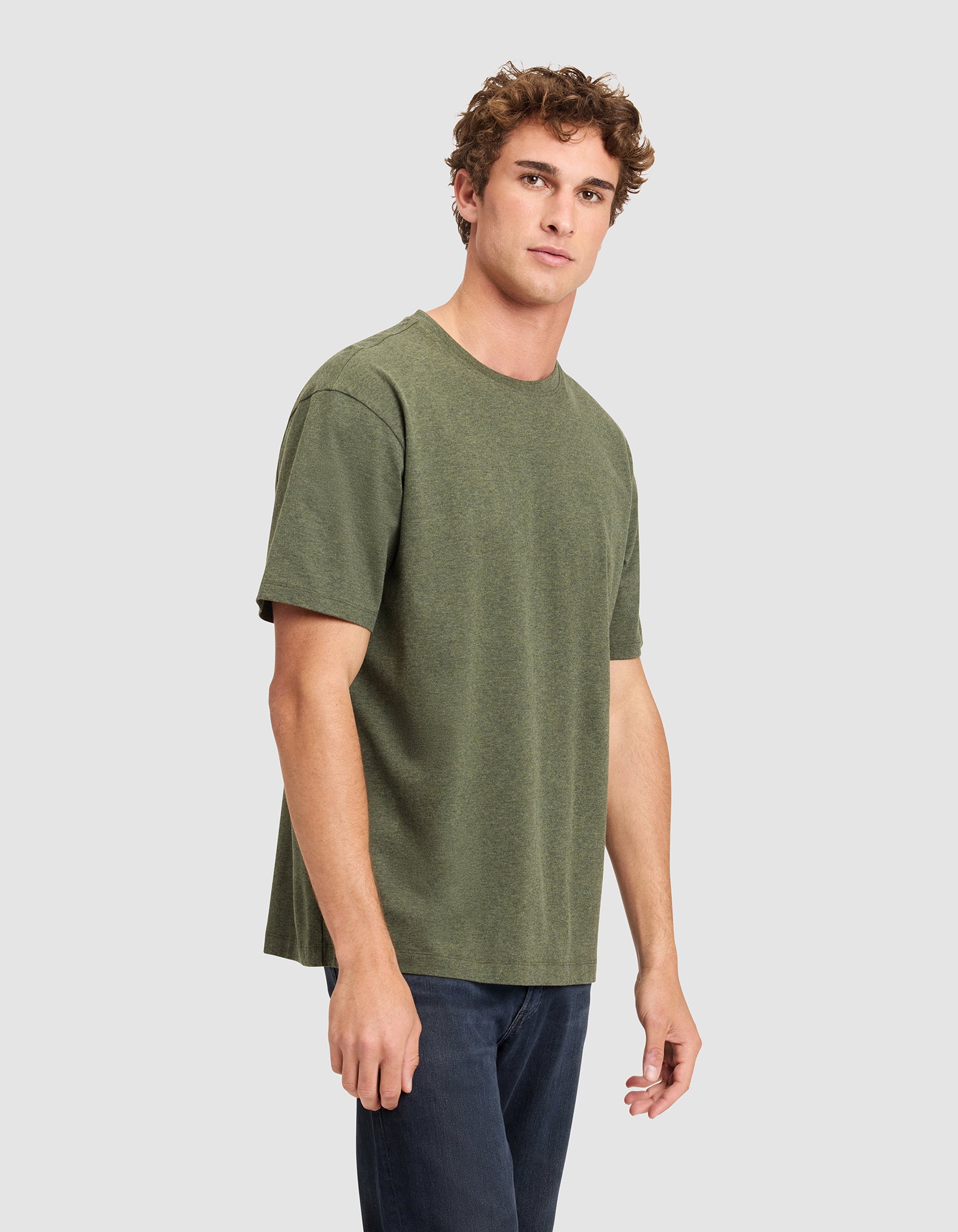 A young man with curly hair wears the Cozy Earth Men's All Day Tee in olive green and dark jeans, standing against a light gray background and gazing slightly to the side. 