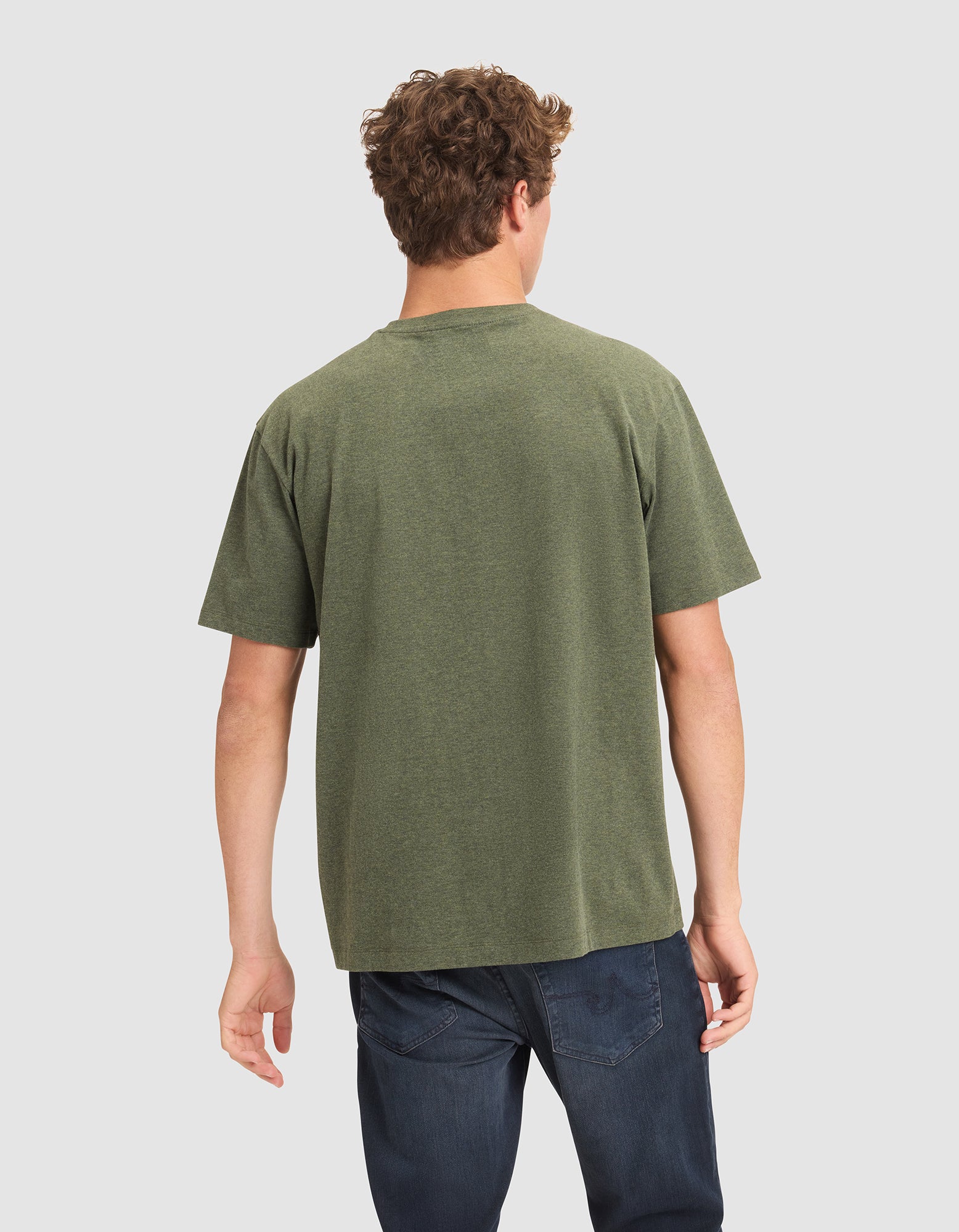 A man with curly hair stands with his back to the camera, wearing Cozy Earth's Men's All Day Tee in olive green and dark blue jeans against a plain light background. 