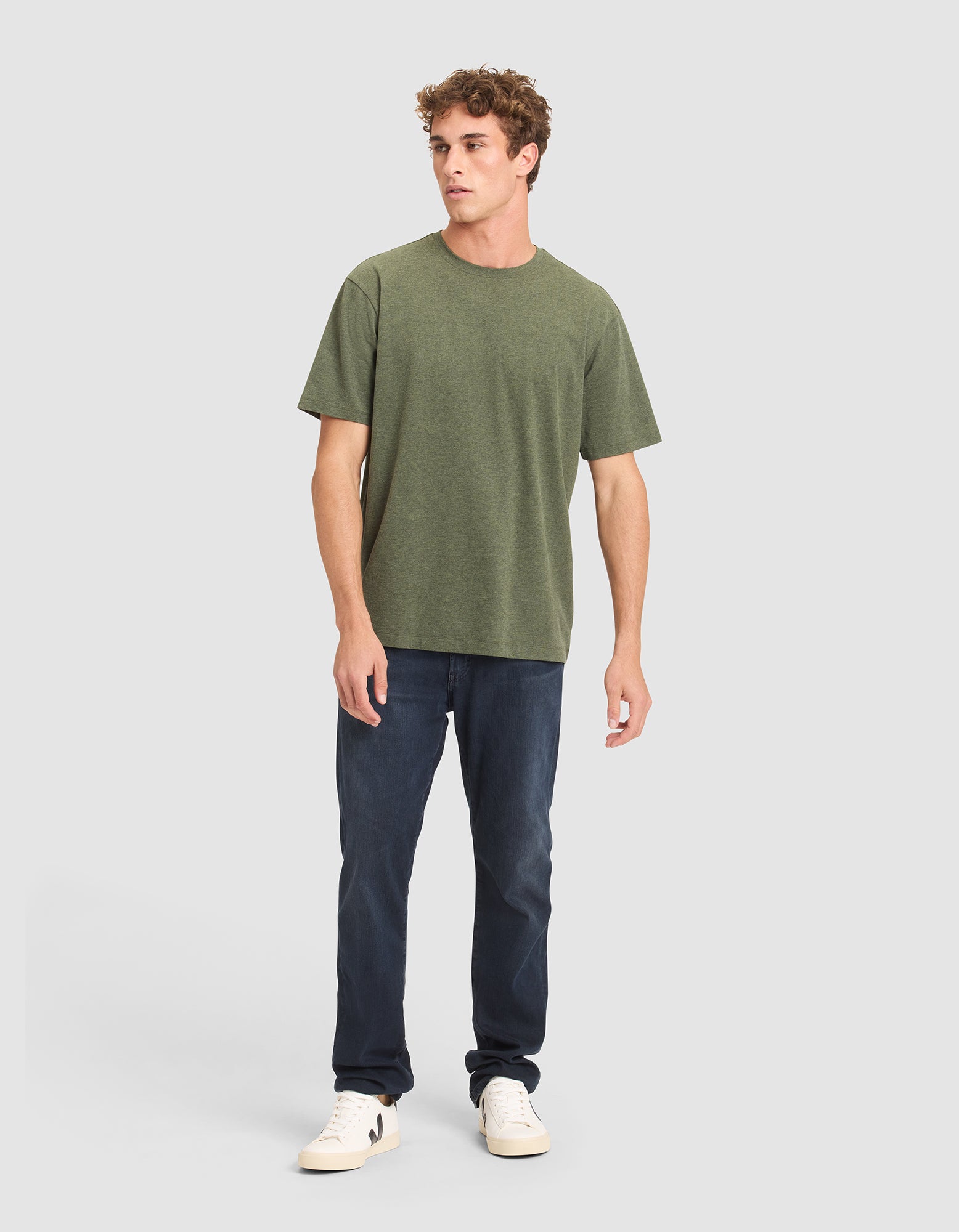 A man with short curly hair and a neutral expression wears the Cozy Earth Men's All Day Tee in olive green, paired with dark blue jeans and white sneakers, standing against a plain light gray background with his left hand relaxed by his side. 