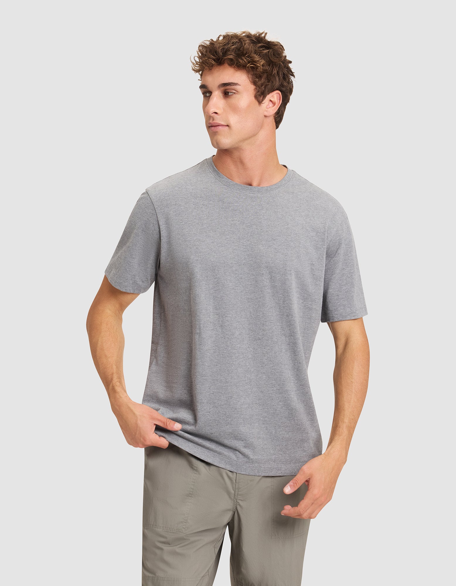 A young man with curly hair wears the Cozy Earth Men's All Day Tee in grey and light khaki pants, standing against a white background with one hand in his pocket and the other adjusting his shirt. 