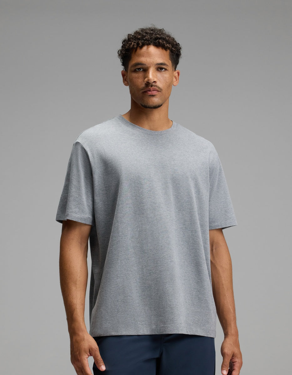A man with short curly hair, wearing the Cozy Earth Men’s All Day Tee in Stone Heather and dark pants, stands against a plain gray background, looking directly at the camera with a neutral expression.