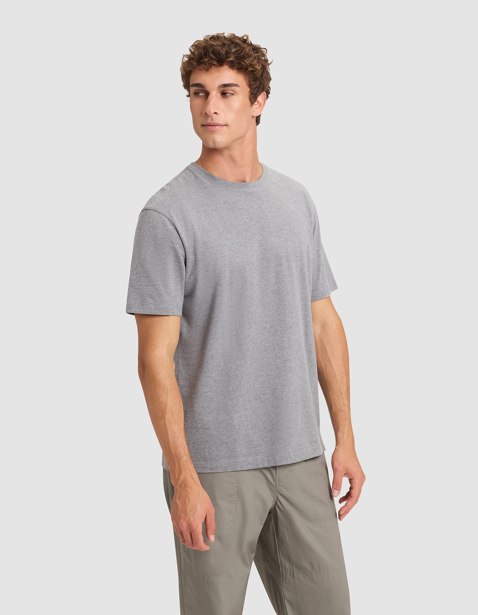 A young man with curly brown hair models the Cozy Earth Men's All Day Tee, a plain grey short-sleeve shirt, paired with light grey pants against a light grey background, gazing slightly left with a neutral expression. 