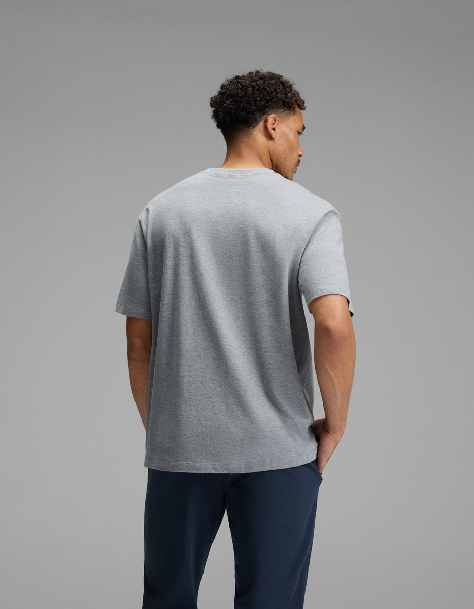 A man with short curly hair stands facing away from the camera, wearing the Cozy Earth Men’s All Day Tee in gray and dark blue pants against a plain gray background.