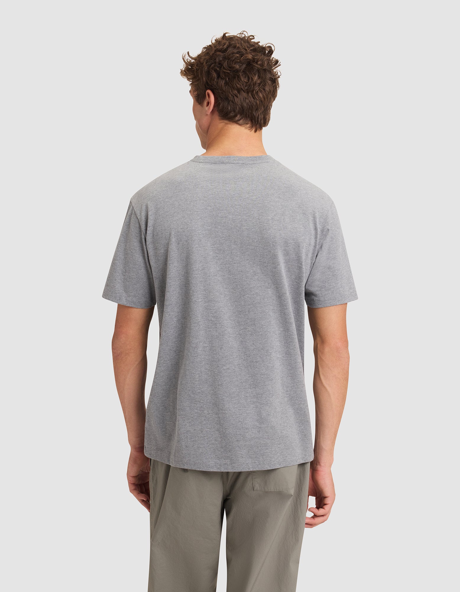 A man with short curly hair is seen from behind, wearing Cozy Earth's Men's All Day Tee in gray and light gray pants, standing against a neutral background. 