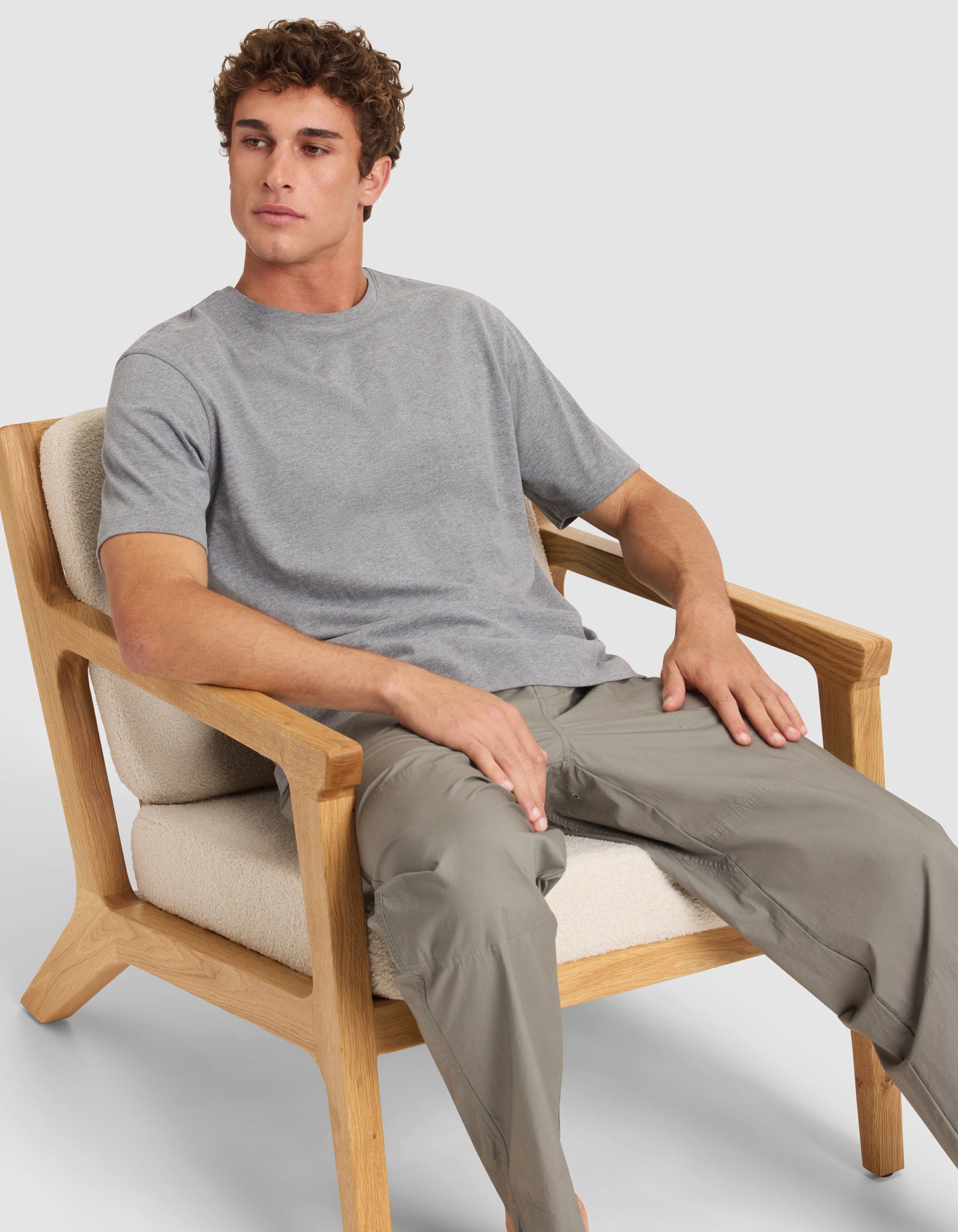 A young man in Cozy Earth's Men's All Day Tee and light gray pants sits in a wooden armchair with cream cushions, looking slightly left with a neutral expression. 