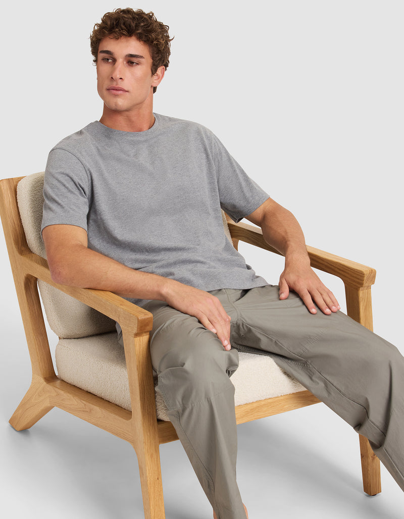 A young man in Cozy Earth's Men's All Day Tee and light gray pants sits in a wooden armchair with cream cushions, looking slightly left with a neutral expression.
