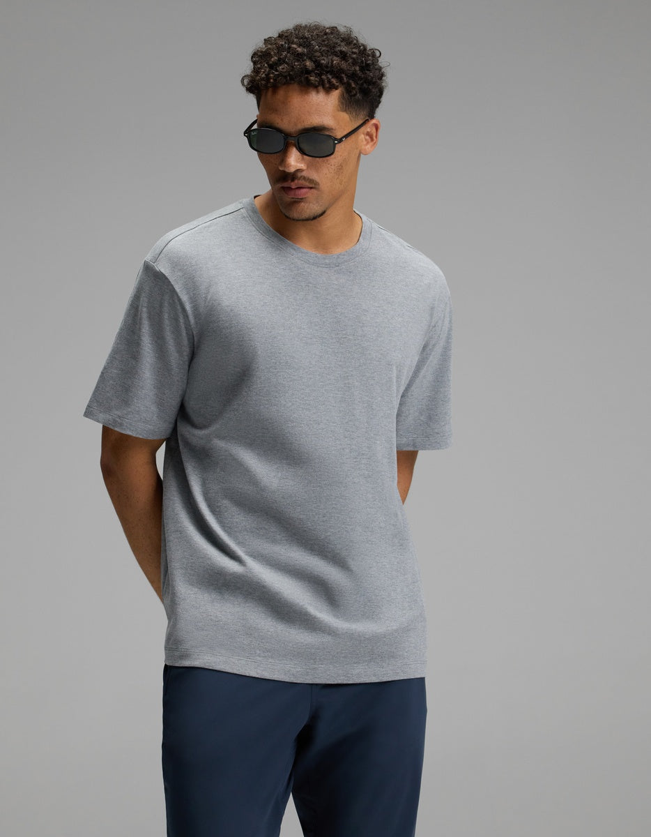 A man stands against a plain gray background with his hands behind his back, wearing the Cozy Earth Men’s All Day Tee in light gray, along with dark blue pants and sunglasses.
