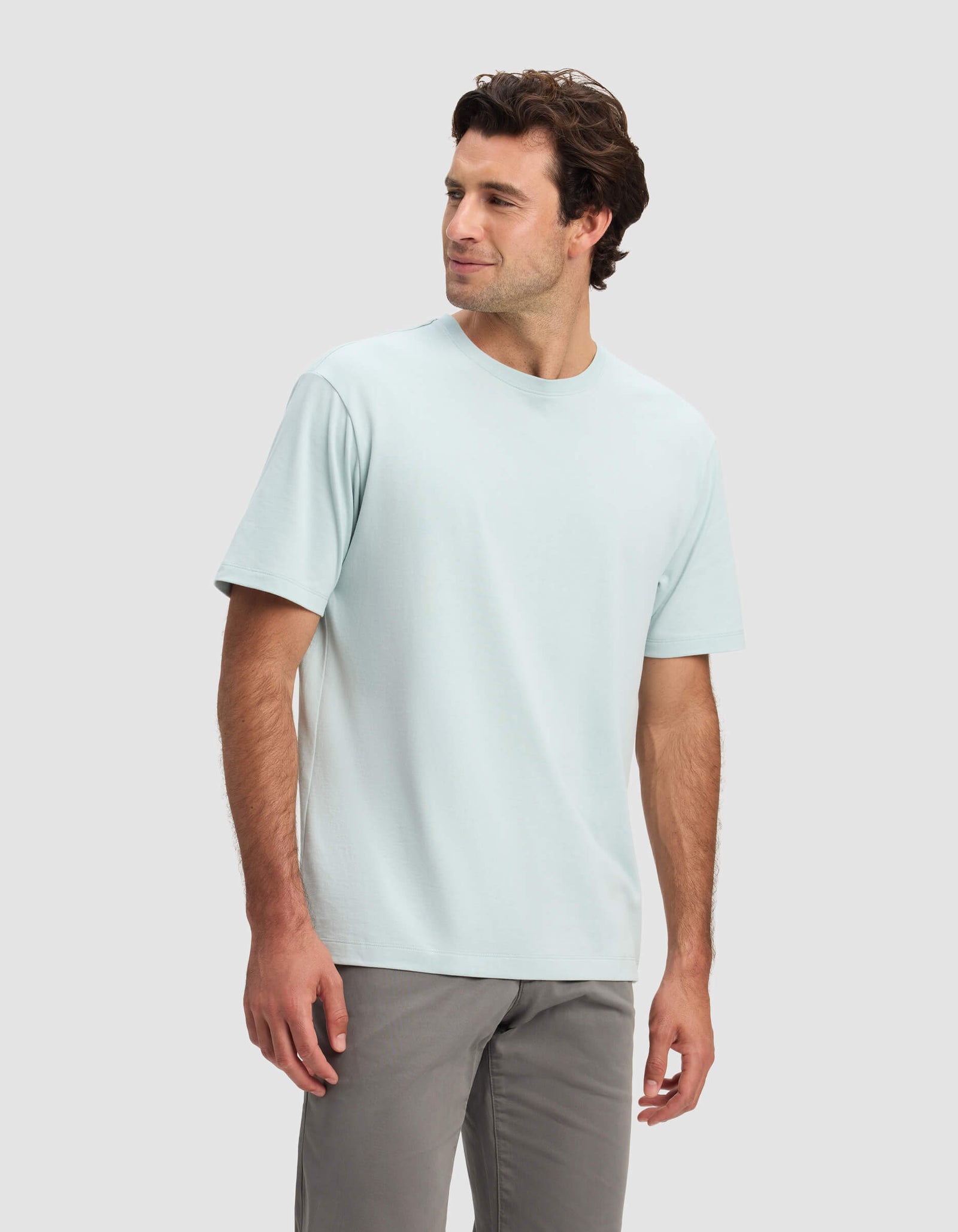 A man with dark hair models the Cozy Earth Men's All Day Tee in light blue and gray pants, standing against a plain light gray background, looking slightly to his left and smiling.