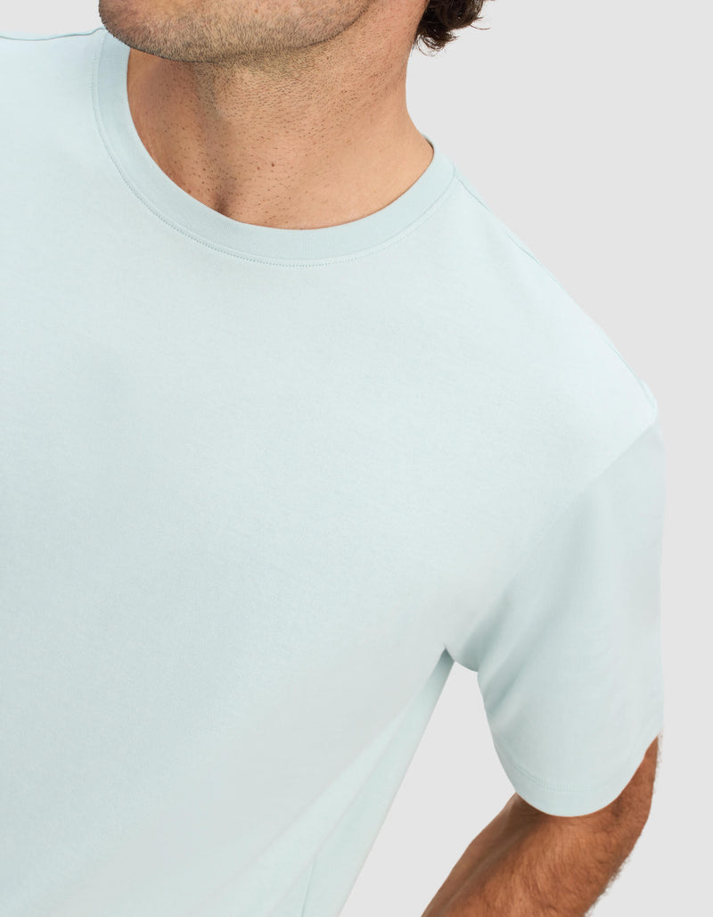 A man is shown from the shoulders up, partially visible against a plain light background, wearing the Cozy Earth Men's All Day Tee in light blue.