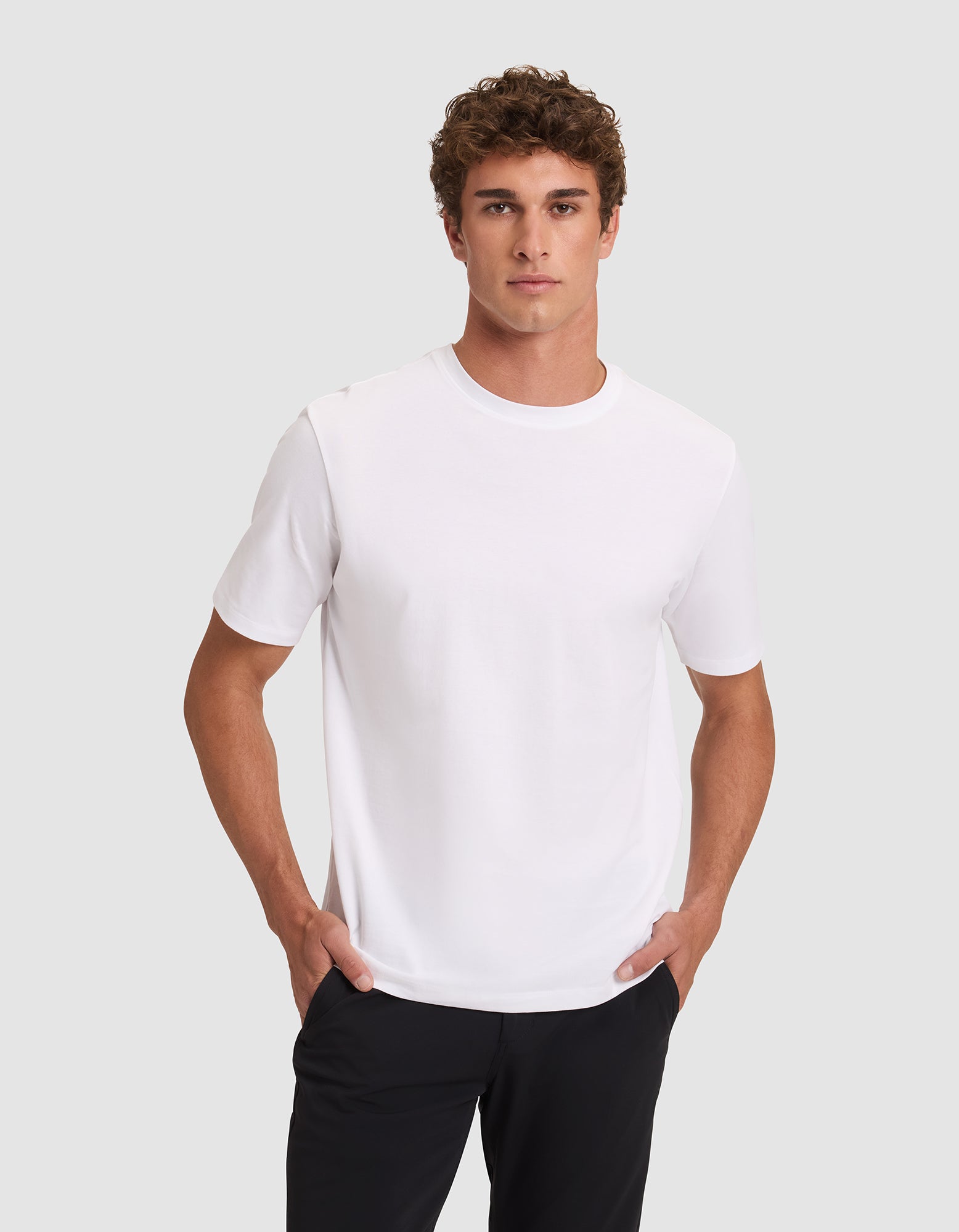 A man with short curly hair wears the Men's All Day Tee by Cozy Earth in white, paired with black pants, standing against a light gray background with his hands in his pockets and looking at the camera neutrally. 